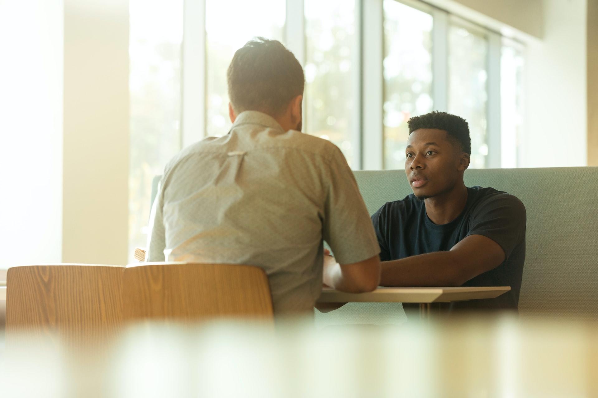 Two men sit across the table from one another, talking. 