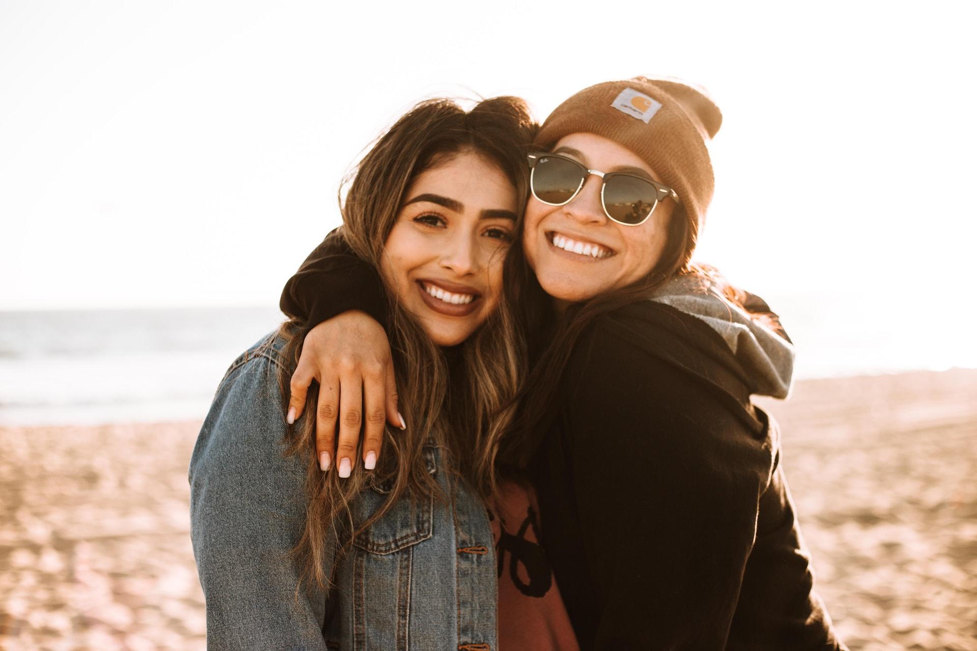 Two women wearing winter gear stand together, arms around one another on a sunny beach.