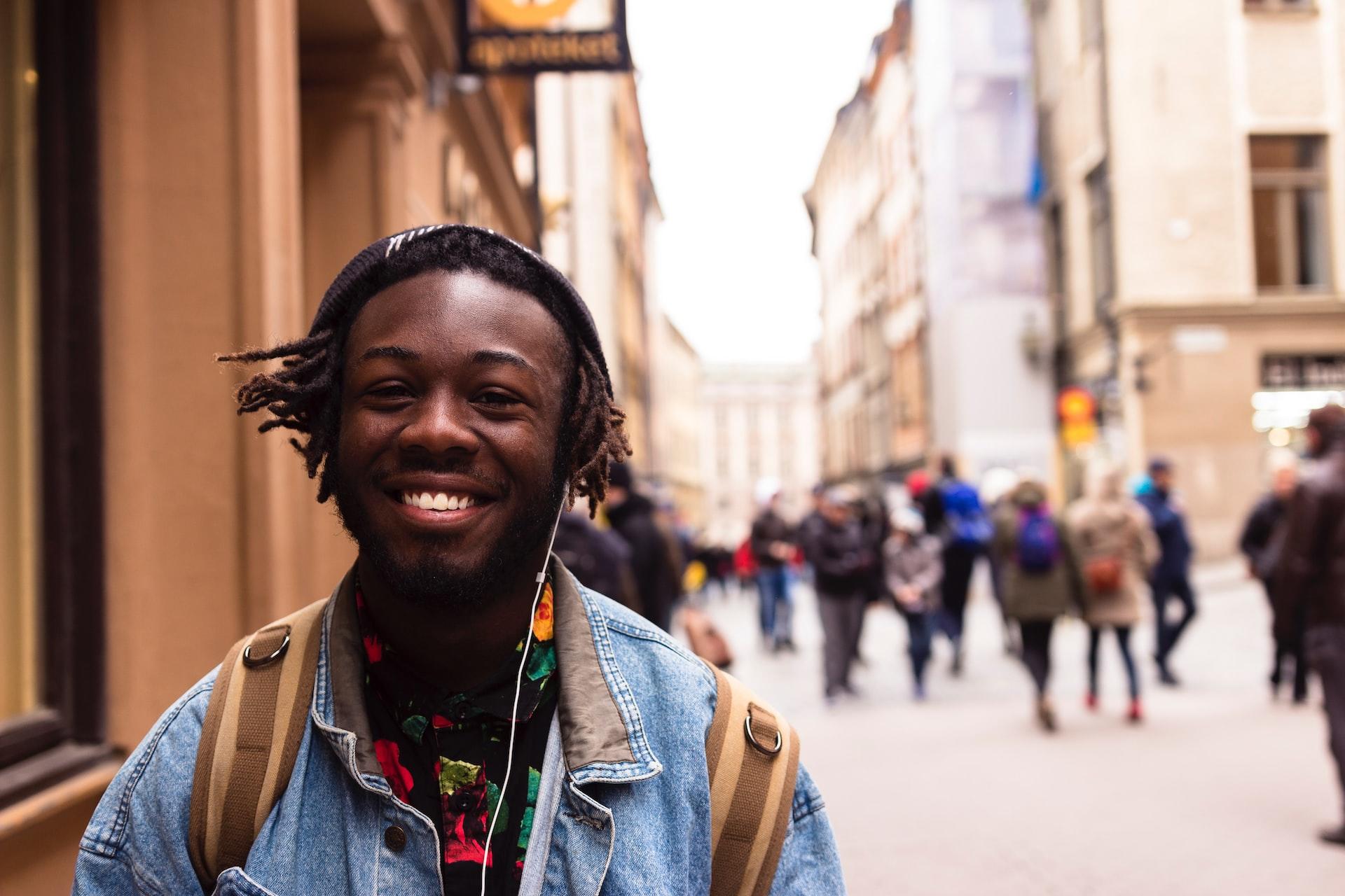 A young man wearing a jeans jacket and carrying a backpack with earbuds in his ears smiles as he walks down the street. 