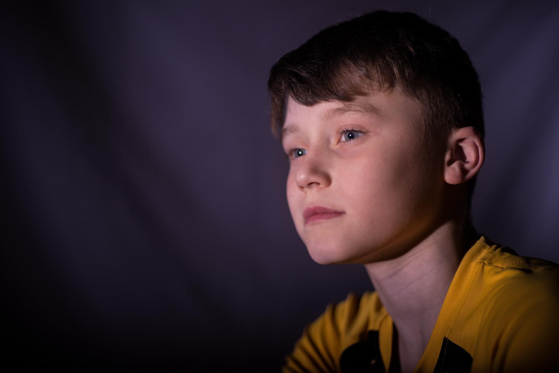 A boy with red hair wearing a yellow and blue shirt appears pensive and unsmiling. 