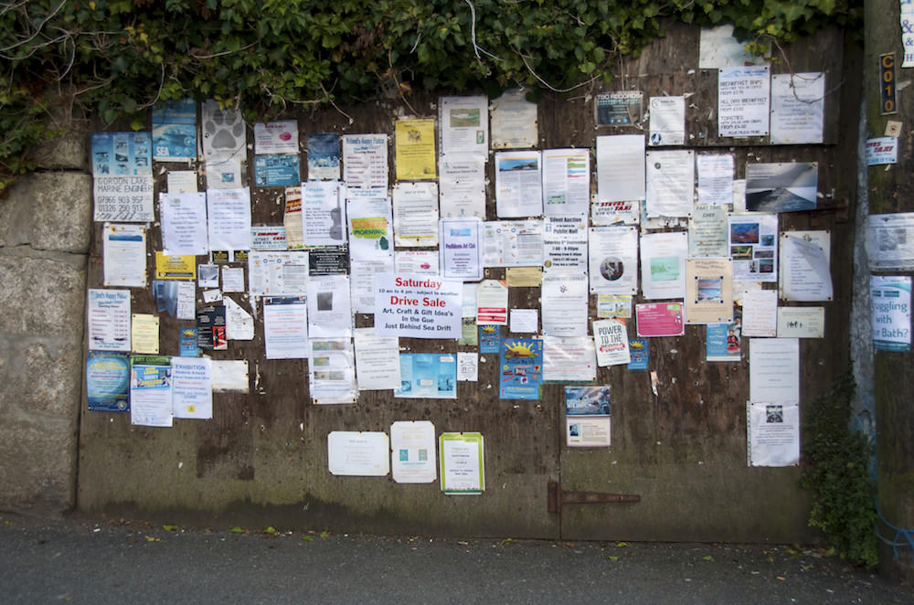 A concrete wall displaying a section of posted notes, adverts and fliers, all in different colours with the majority of them being white paper. 