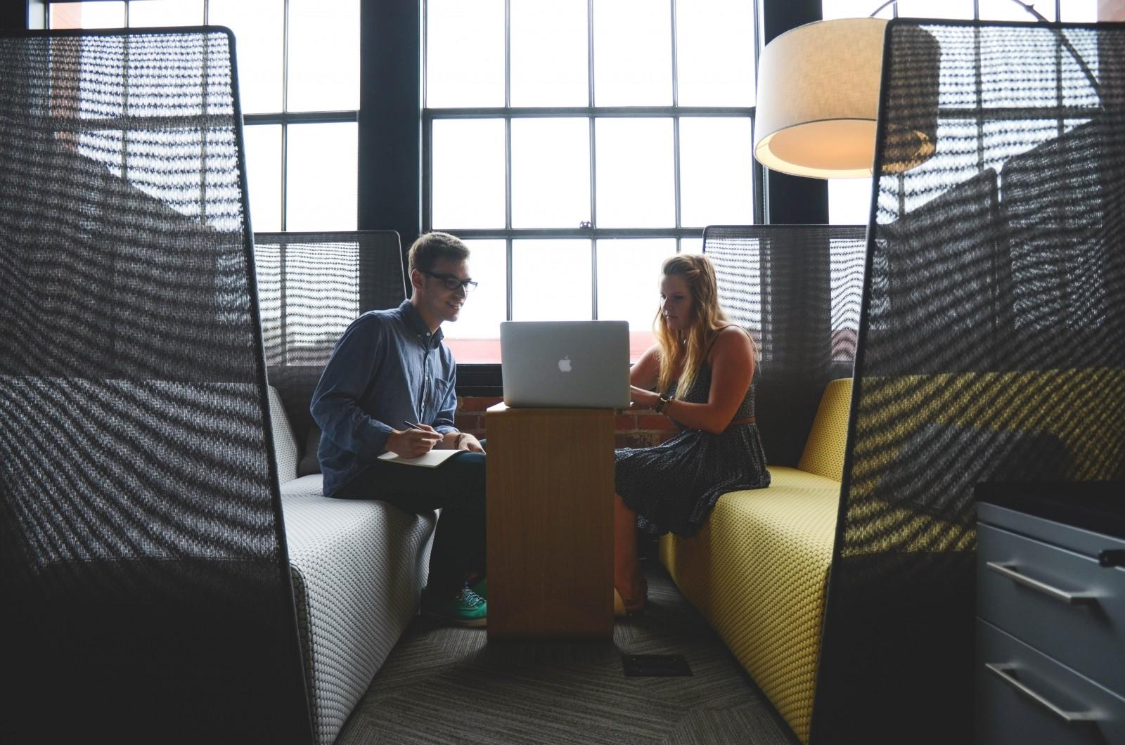 Two people sit across from one another with a narrow wooden table separating them, upon which rests an open laptop computer whose screen faces away from the camera. 