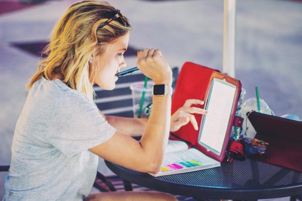 A person wearing a grey top and shorts sits at an outdoor table with a pen in their right hand while their left hand glides over their tablet screen, which they have propped up against the table's umbrella post.
