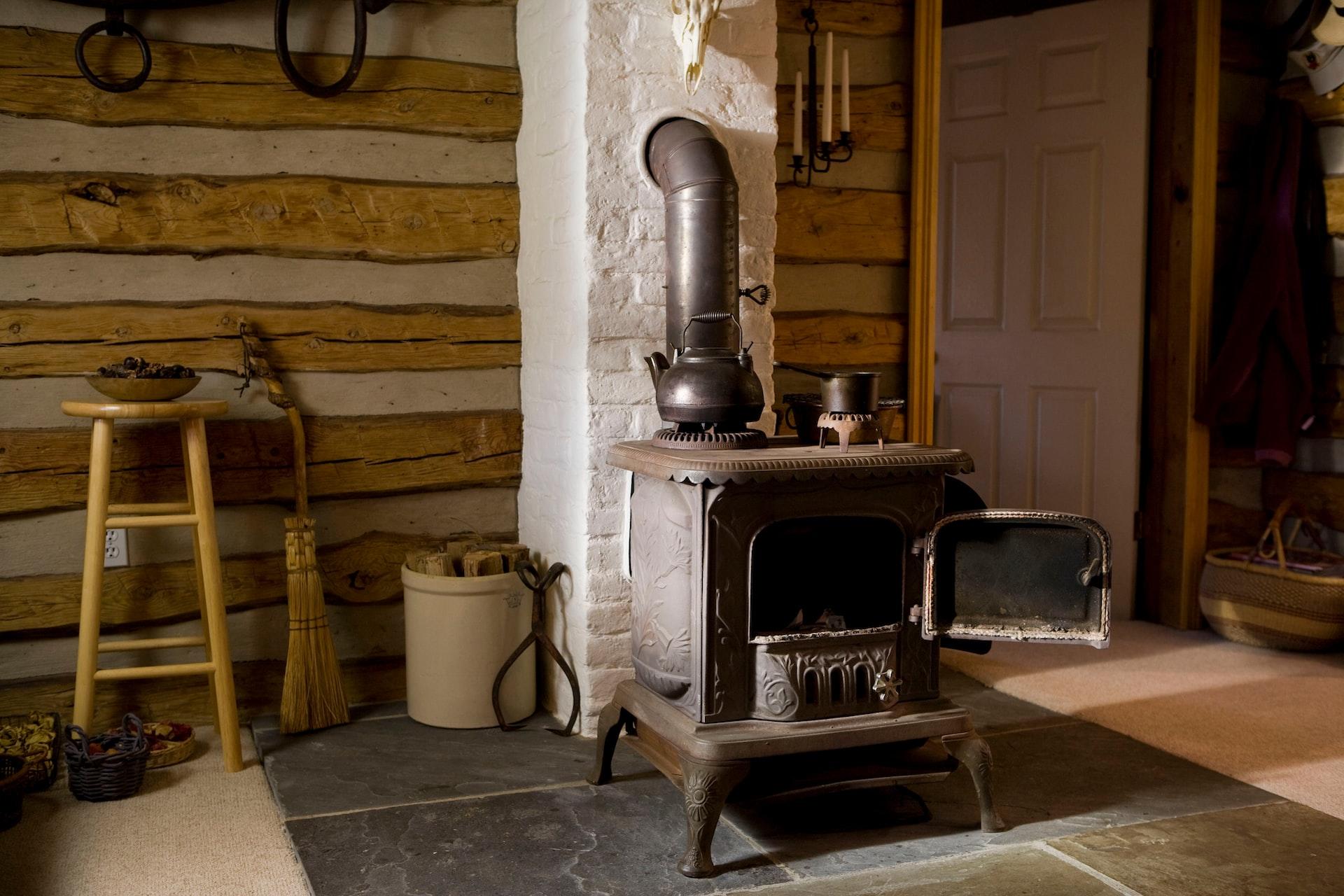 A wood-burning stove set in front of a chimney column in front of a wood beam wall with hearth tools leaning against it.