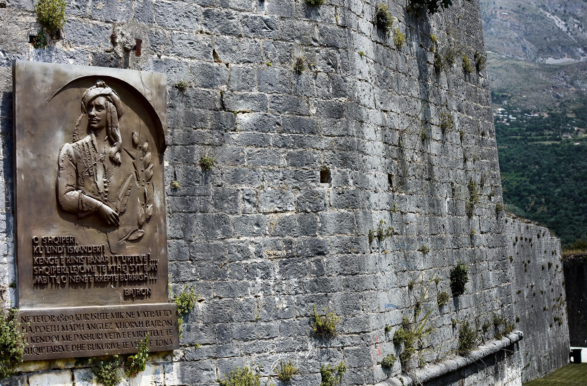 A city wall in Albania bearing a bronze plaque dedicated to Lord Byron with an expanse of greenery visible in the background.