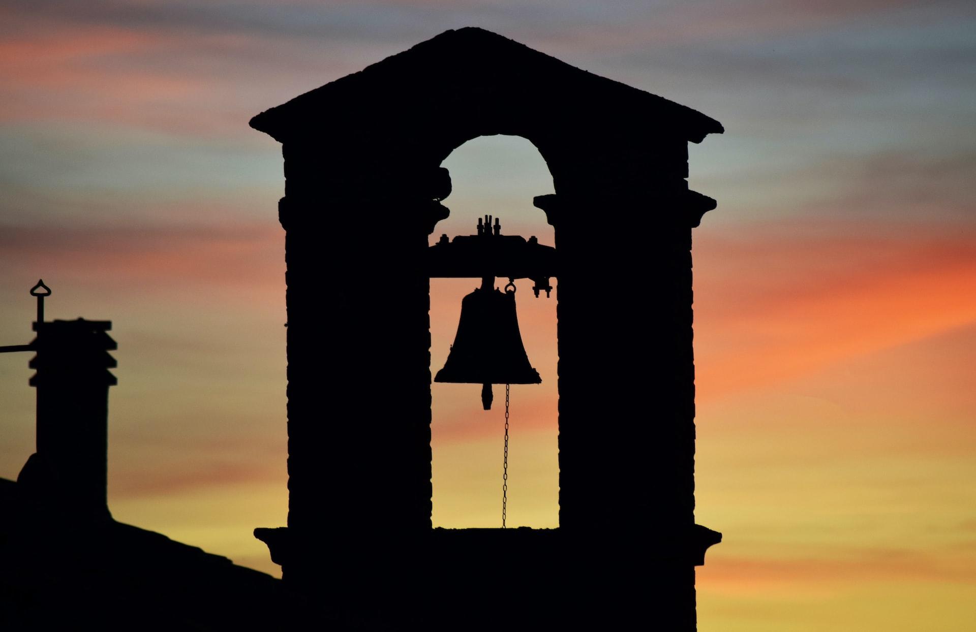 A bell mounted in an arched structure over a roofline seen in contrast with the twilight sky of orange, yellow and lavender. 