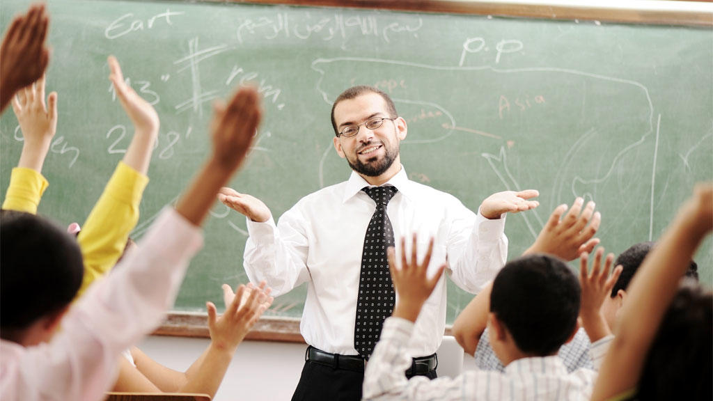 A teacher wearing a white shirt and tie stands in front of the chalkboard and faces their classroom, where the students all have their hands raised.