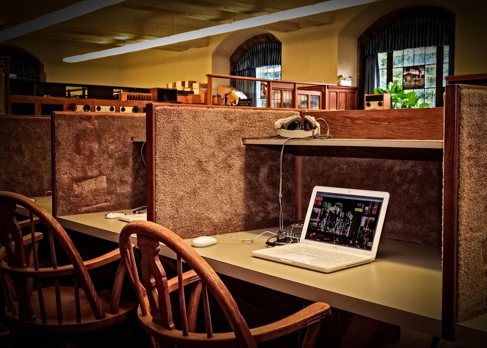 One cubicle in a study lab that features carpeted walls and a shelf. An open laptop sits on the writing surface and a bentwood chair waits for someone to occupy it.