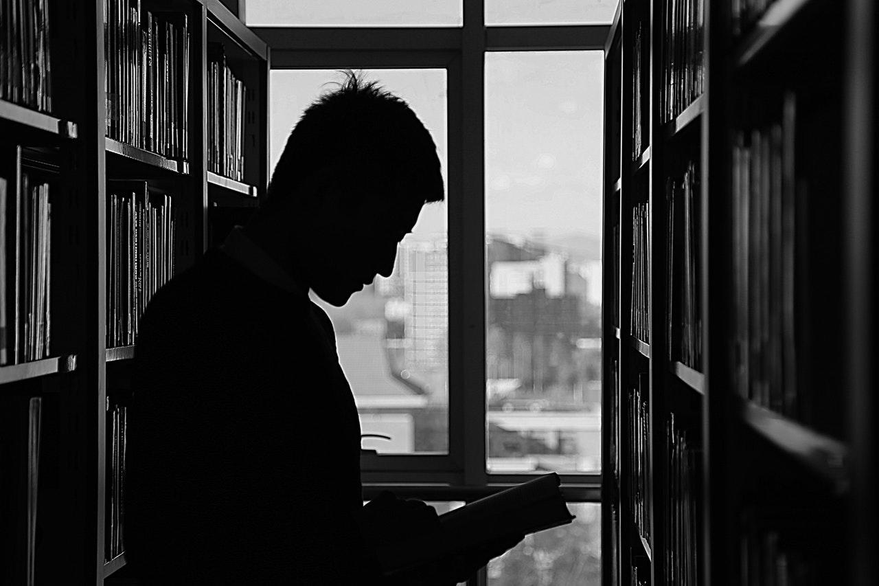 A person seen in silhouette between two library stacks with a book in his hand and a window in the background filtering in natural light.
