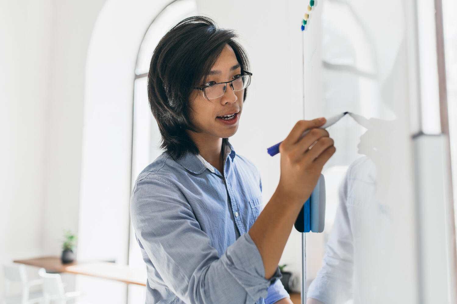 A person in a blue shirt is writing on a whiteboard with a marker in a bright, modern room with large windows.