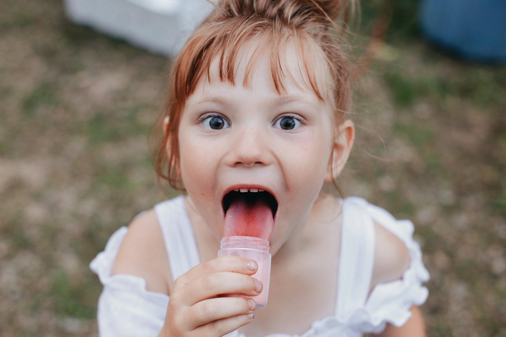 A young girl with red hair and blue eyes wears a white dress and looks directly at the camera while licking a sweet out of a small, clear container.