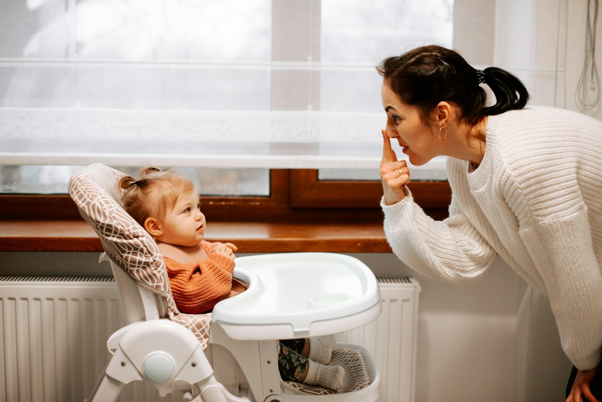 A person with dark hair, wearing a white sweater leans down toward a child in a high chair, pointing at their nose.