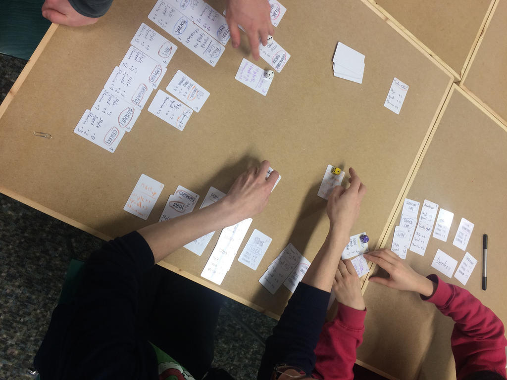 Three people playing a word game using white index cards against a brown table, seen from overhead.
