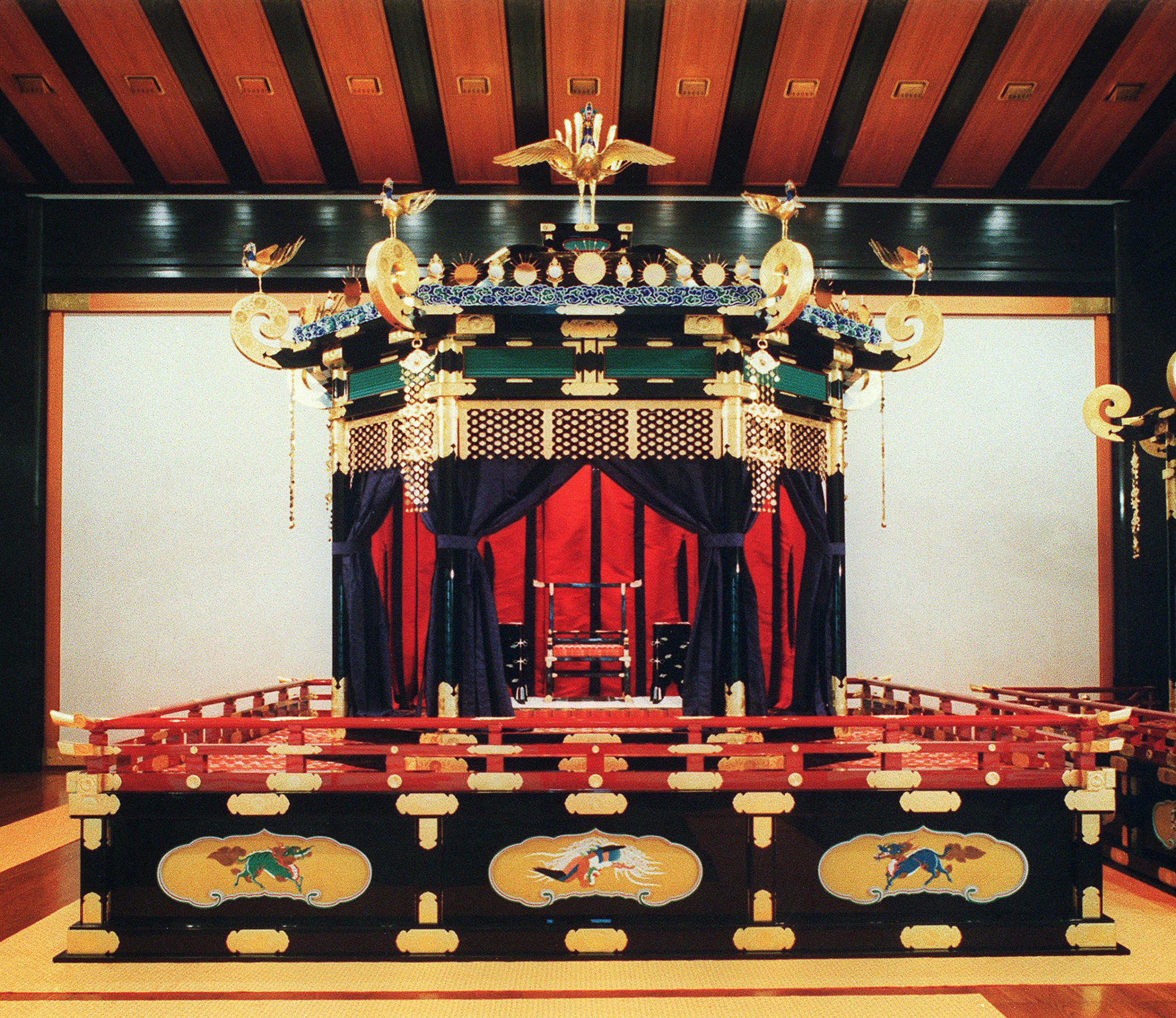 A Japanese throne displayed in an enclosure of black and red with gold accents, set behind a red, gold and black barrier. 