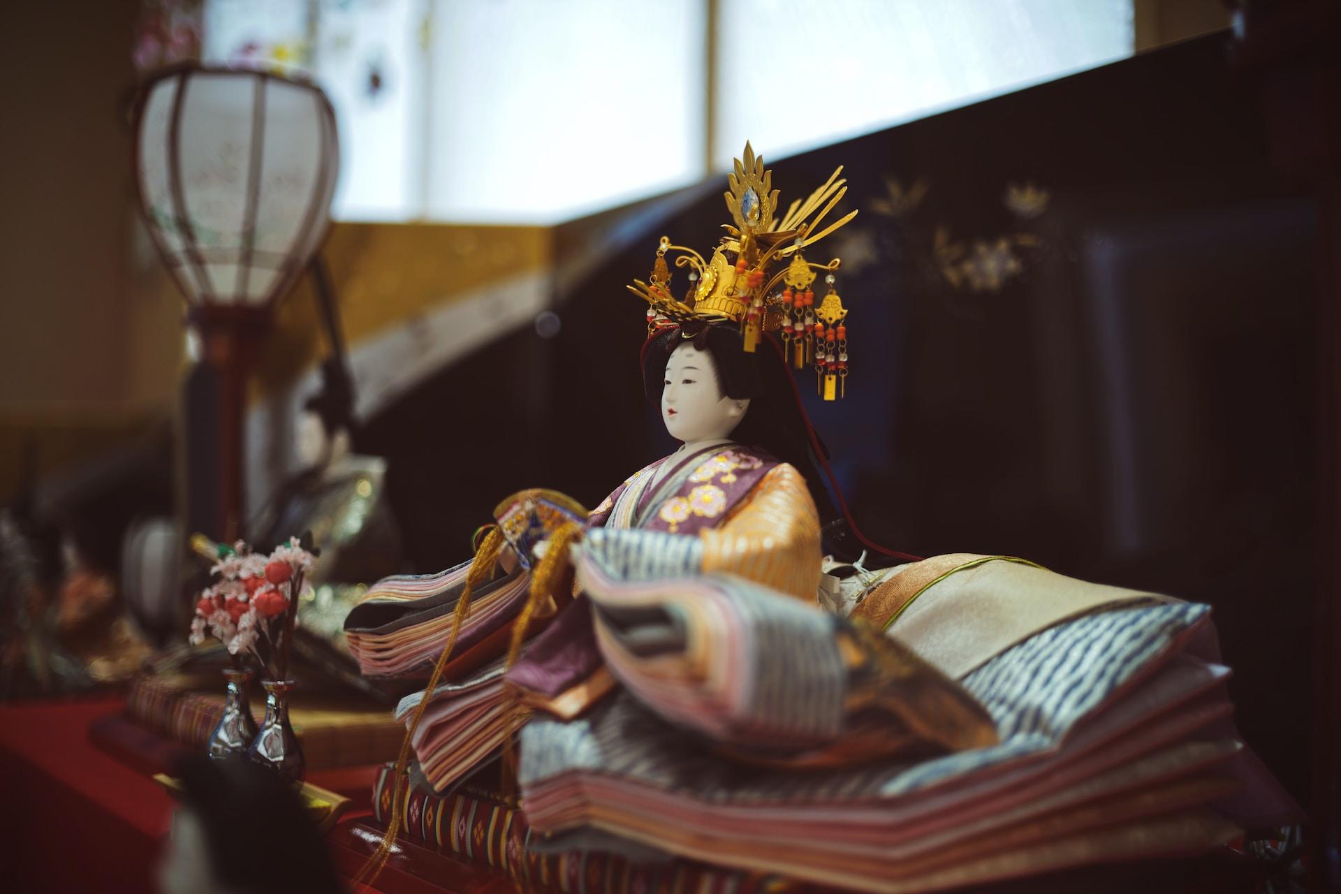 A side view of a geisha doll lavishly dressed and posed on an altar against a dark background. 