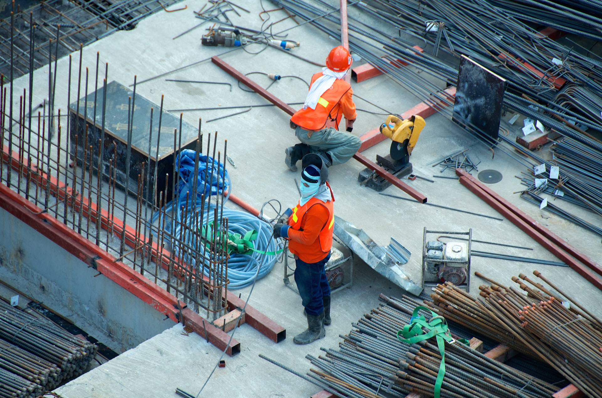 A commercial building site with bundles of rebar laying on a concrete foundation and two workers in orange protective vests preparing to work.