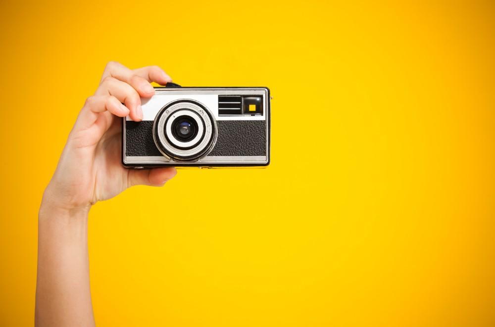 A hand holding up a point-and-shoot camera which has a black and grey body, against a yellow background.