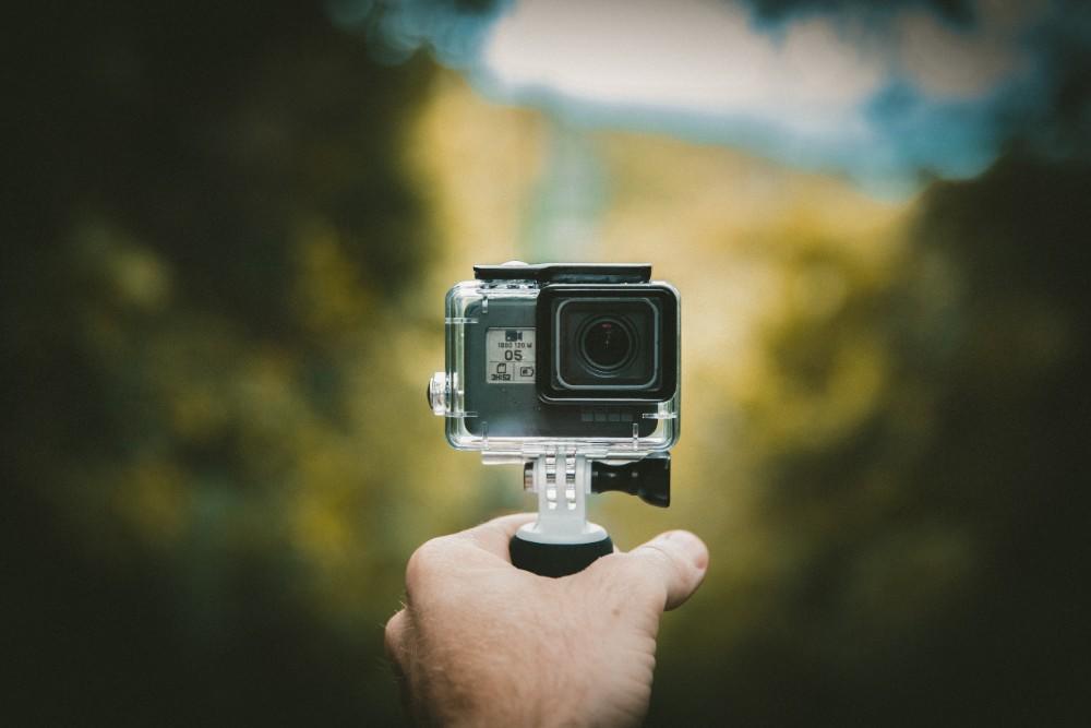 A hand holds a GoPro camera's stem, showing the camera's face in sharp contrast with the blurred background, which is dark green at the edges and lighter yellow towards the centre of the frame.