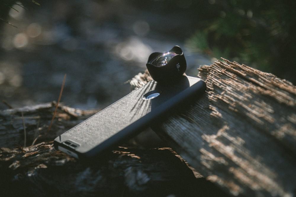 A device resembling a smartphone with an added camera lens attached, sheathed in a black case, rests on the ground, propped on a piece of wood. The sun shines on it.