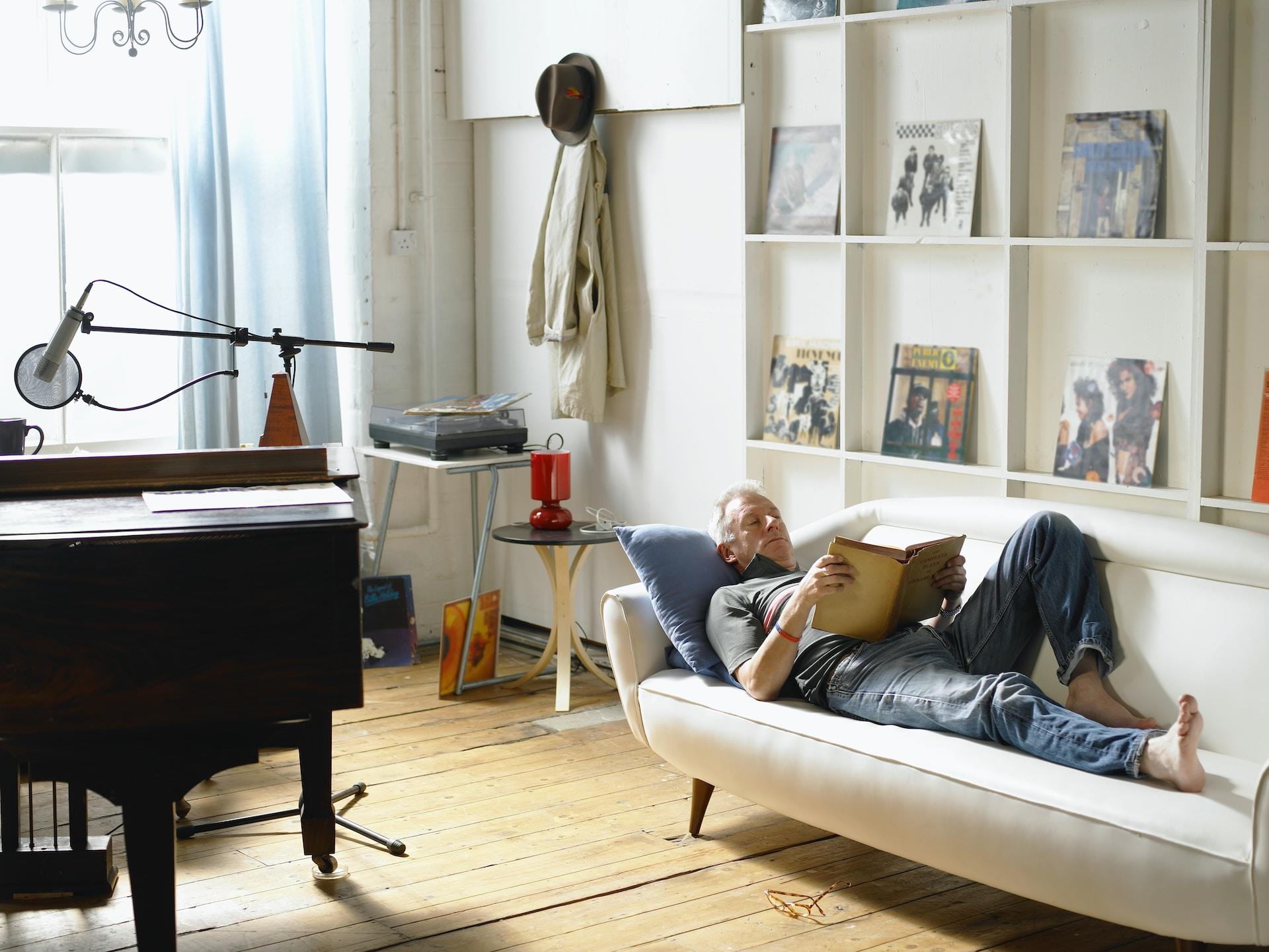Man is laying in a sofa, next to a piano, reading a book.