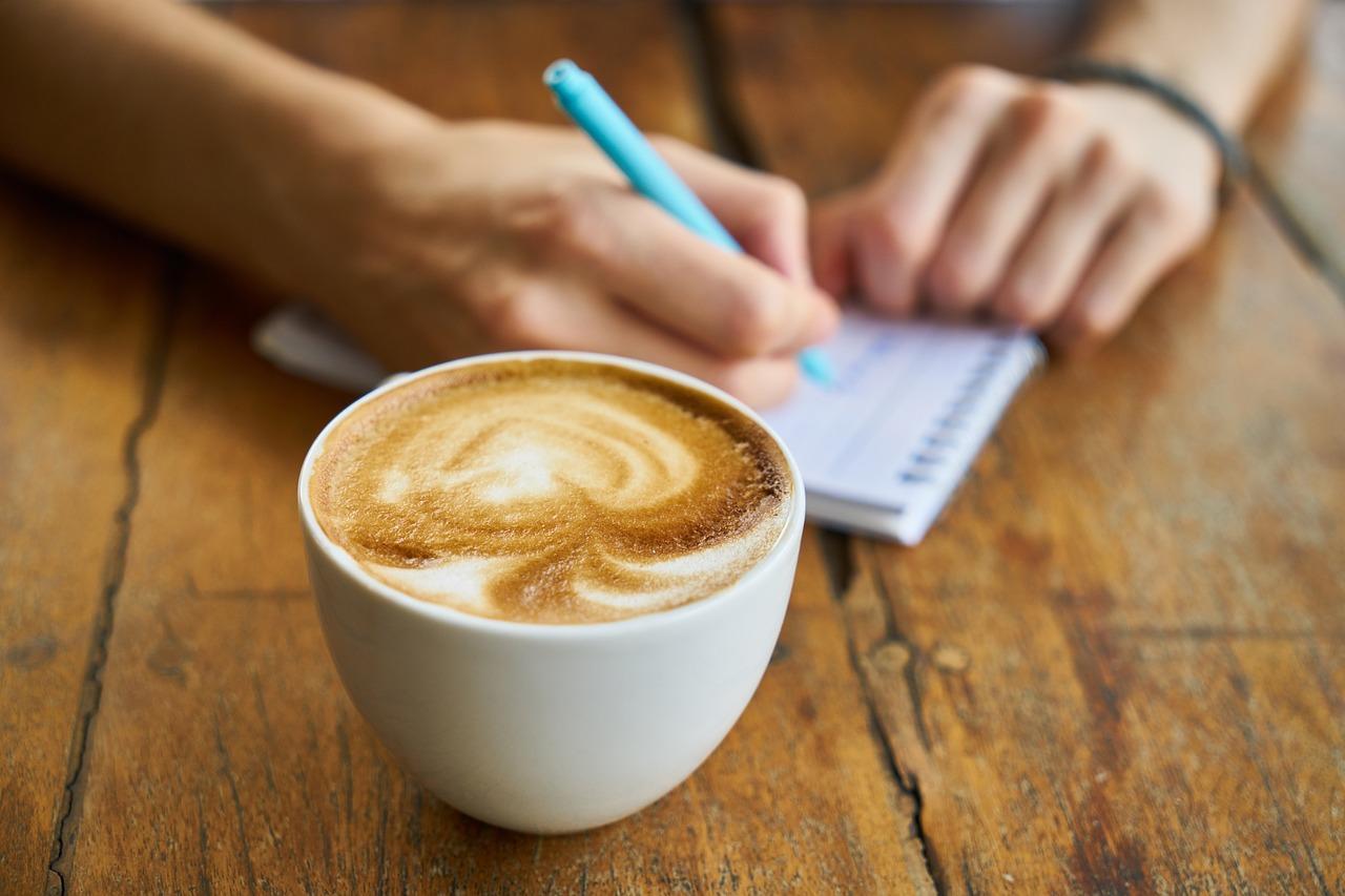 An image of someone's hands writing something down on a notepad with a coffee in a mug next to them. Learning Portuguese often means learning how to write in Portuguese as well.