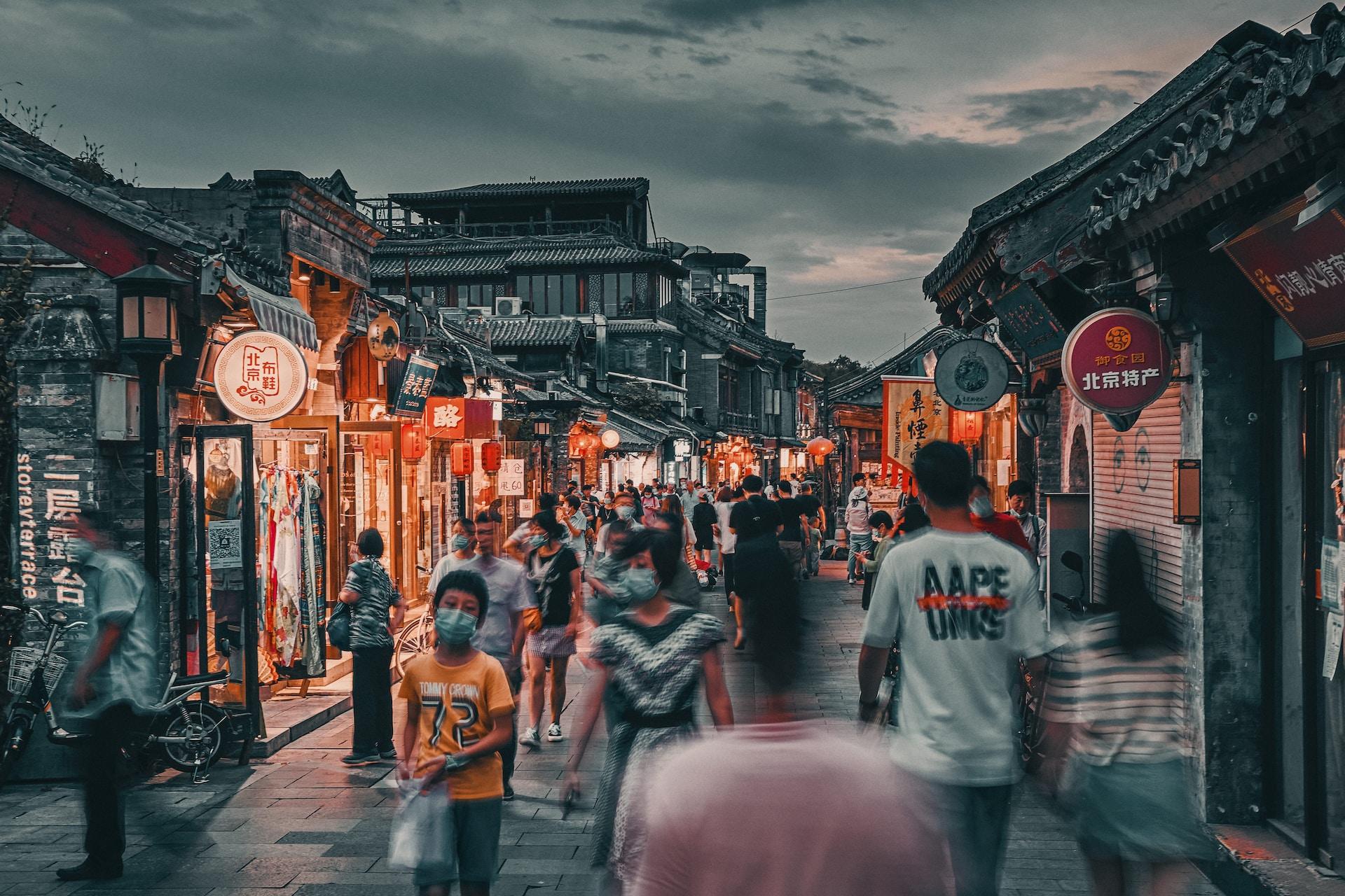 A Beijing walking street filled with pedestrians at dusk, with shopkeepers just starting to turn on their neon signs.