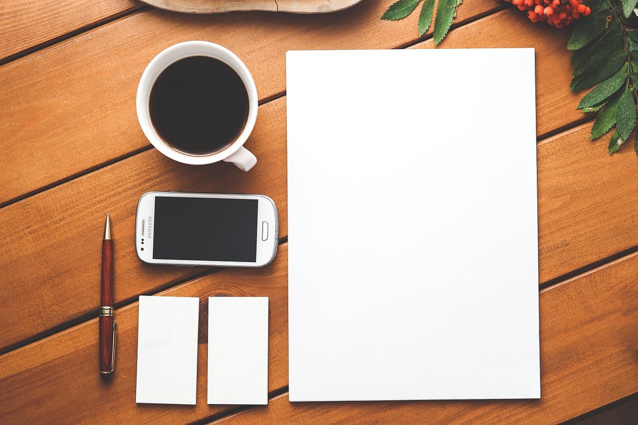 A photo of a pad of white paper, two white pads of small paper, a pen, a mobile phone, and a cup of black coffee in a white mug. Learning business Russian is possible with some Russian classes.
