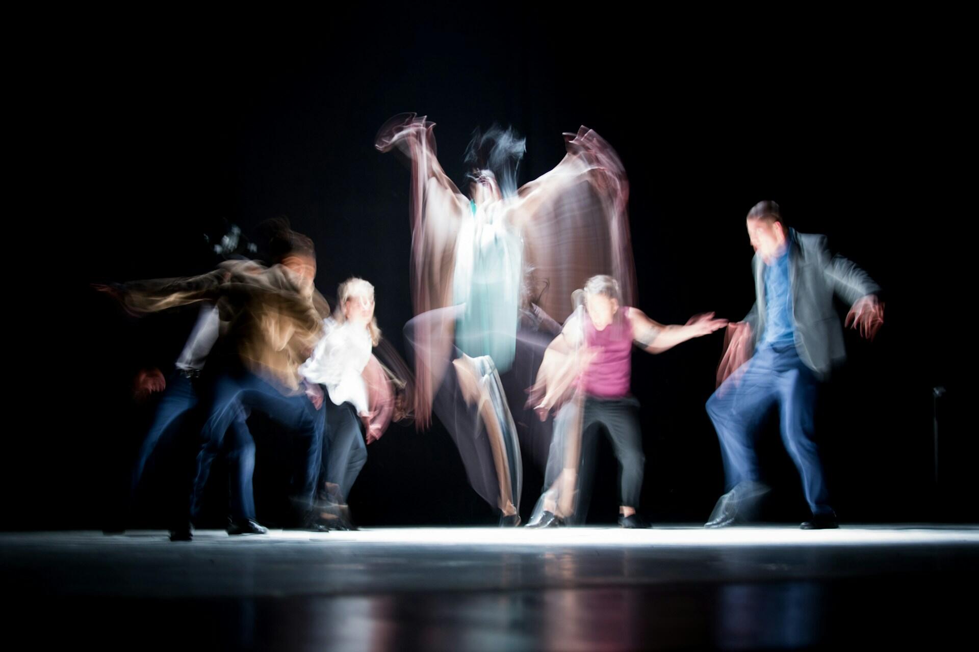 A dynamic group of dancers performs on stage, their movements captured in a blur against a dark background, creating a sense of motion.
