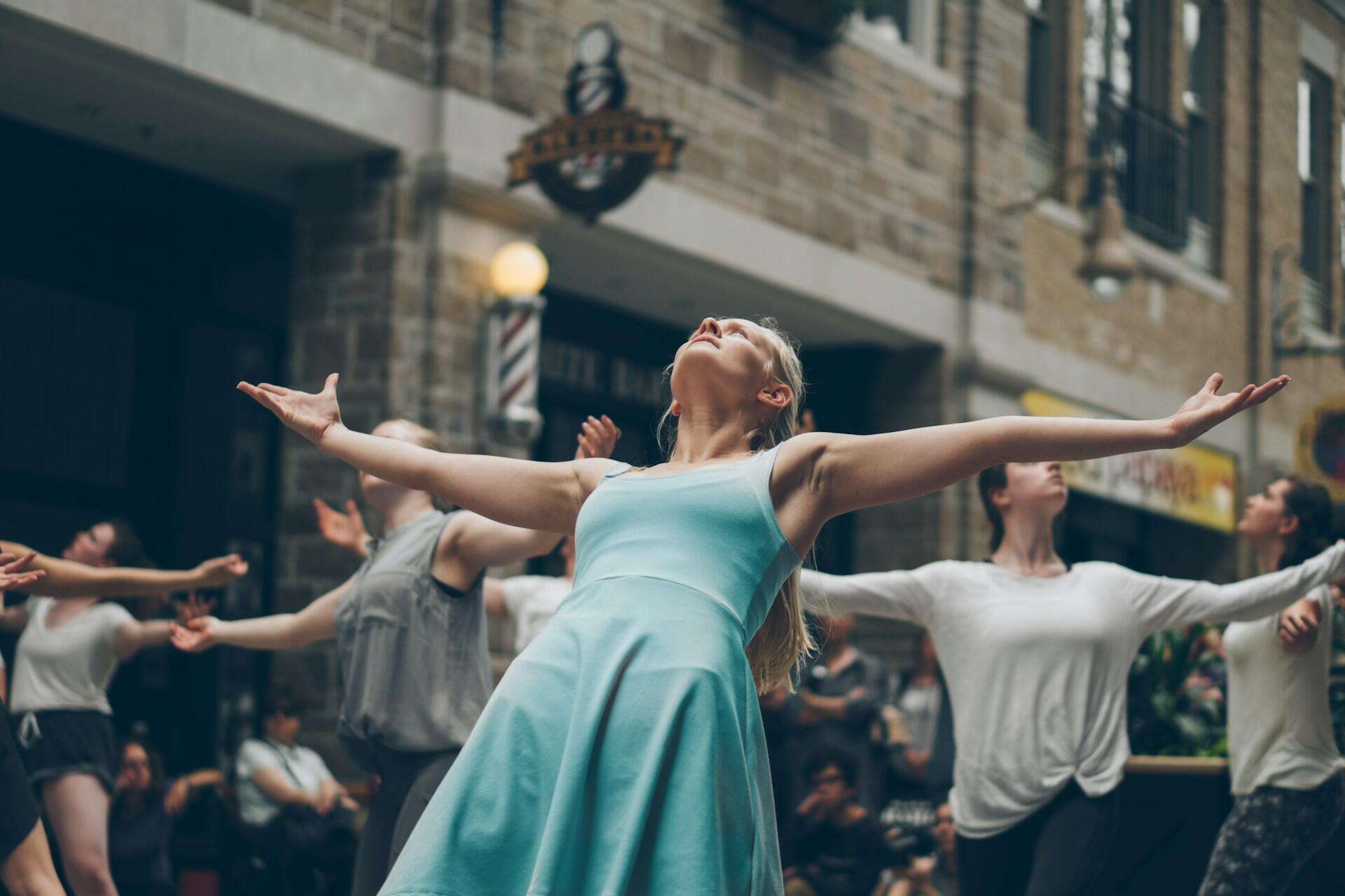 A dancer in a light blue dress performs expressively, surrounded by fellow dancers in a lively, urban setting.