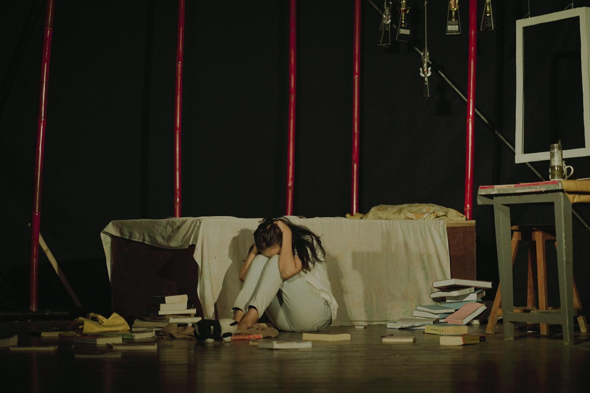 A person sits curled up on the floor, surrounded by scattered books, in a dimly lit room with minimal furnishings.