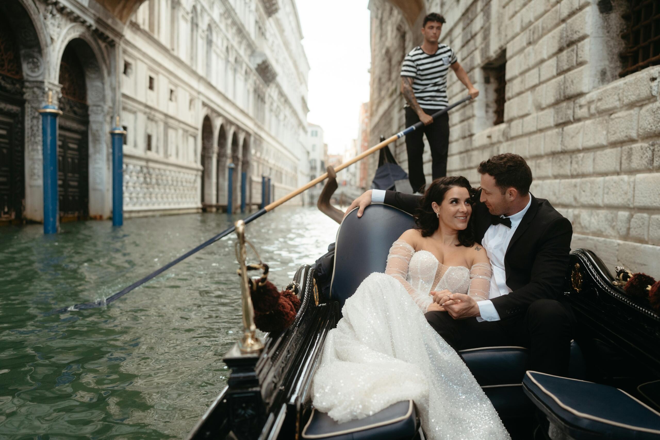 A couple dressed elegantly is enjoying a romantic gondola ride through the serene canals of Venice, surrounded by historic architecture.