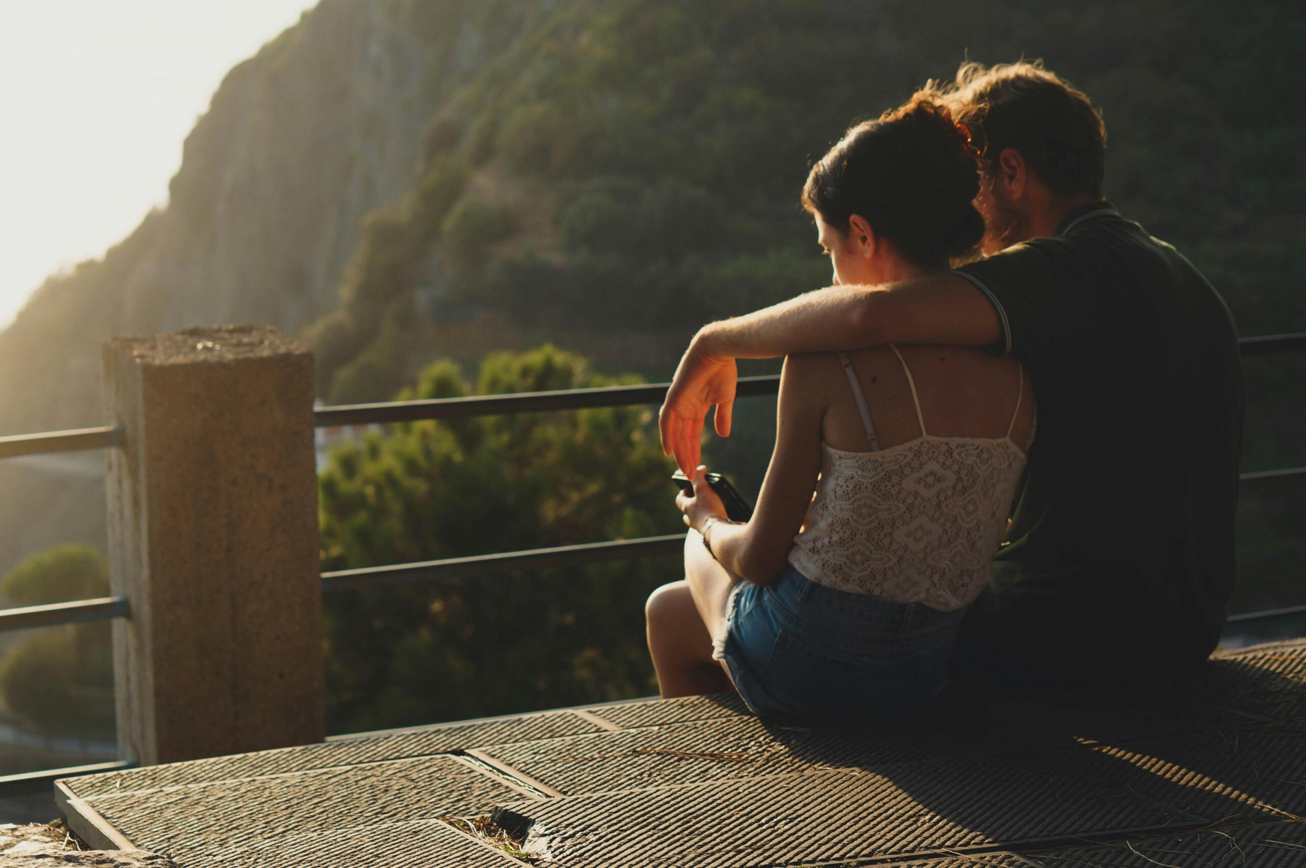 A couple sits closely together on a sunlit platform, with a scenic view of green hills in the background, enjoying a peaceful moment.