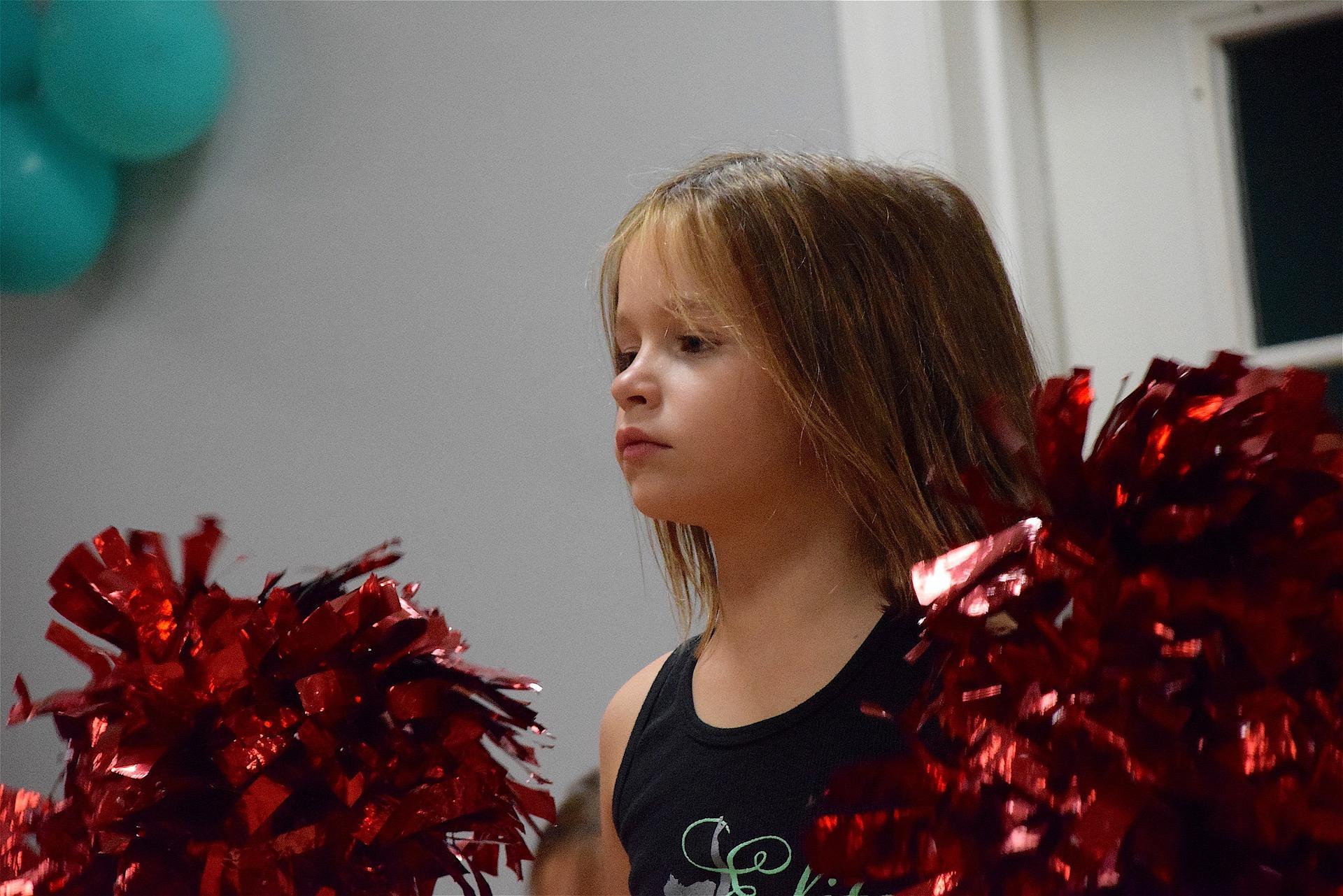 A little girl is wearing a black top and carrying pom-poms while dancing. Dance classes for kids can also prepare the little ones for a future career in cheerleading. Cheerleaders go through immense training and are required to be in peak physical condition – much like athletes