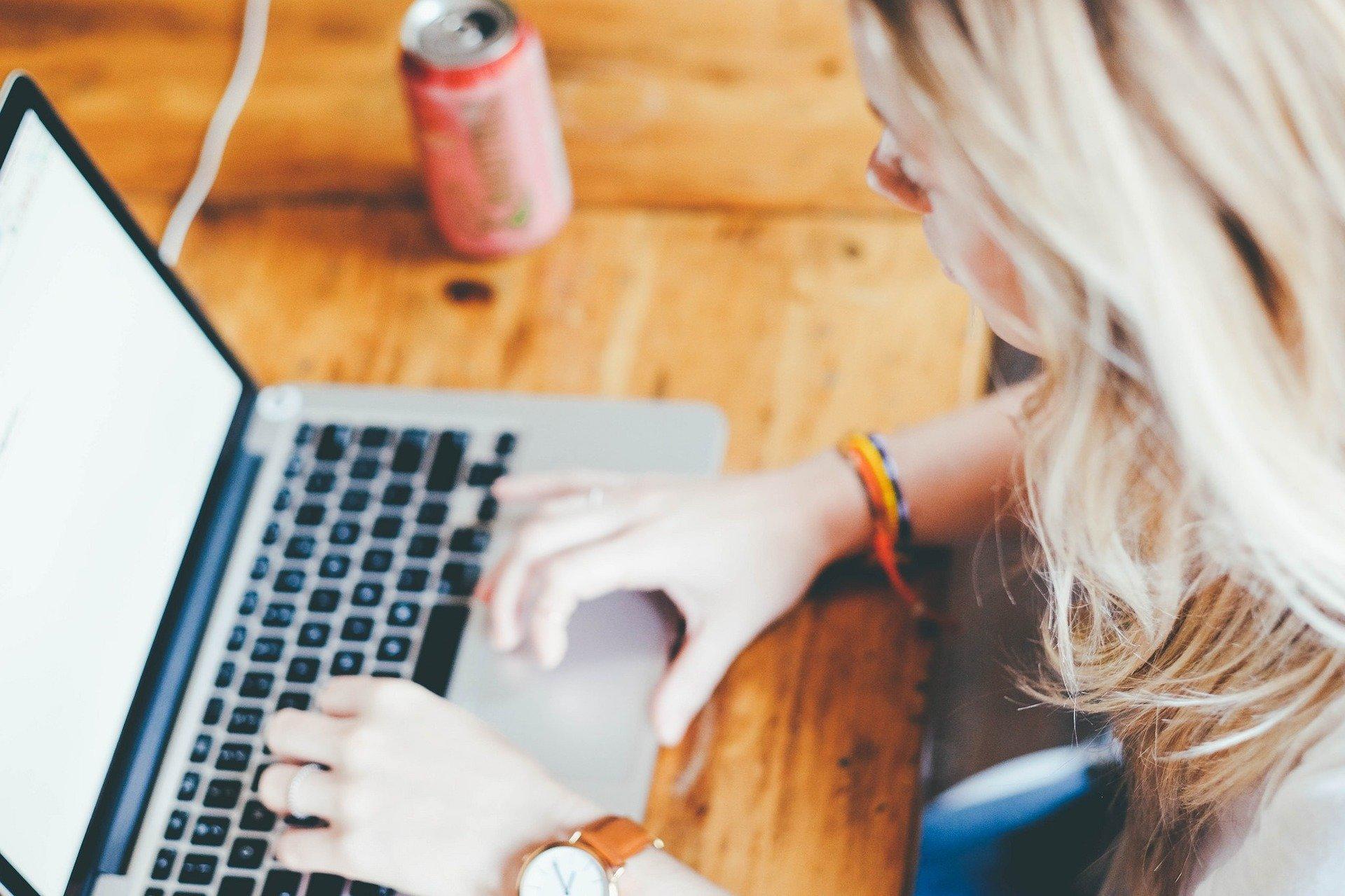 A blonde woman sits at a light-coloured wood table with an open laptop computer, typing on it.