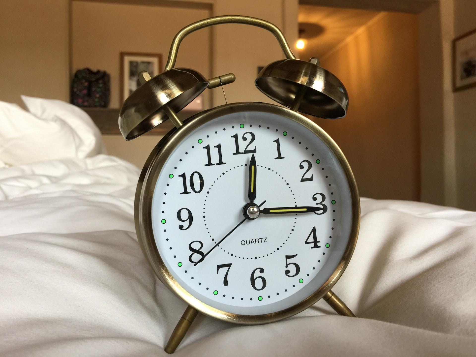 An golden analogue alarm clock with twin bells and a hammer in between them stands upright on a white coloured duvet