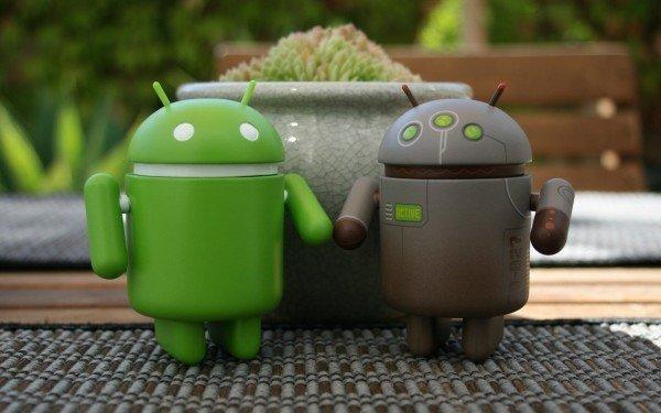 Two android figurines, one green with white markings and the other grey and brown with green markings, stand in front of a flowerpot on a slatted wood table. 