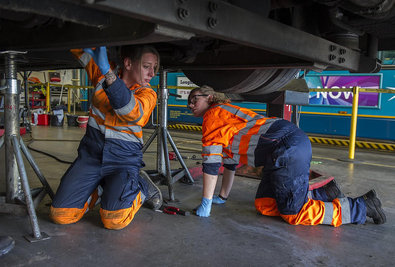 Two women in orange and blue work coveralls, wearing blue nitrile gloves kneel under a vehicle set up on jack stands. 