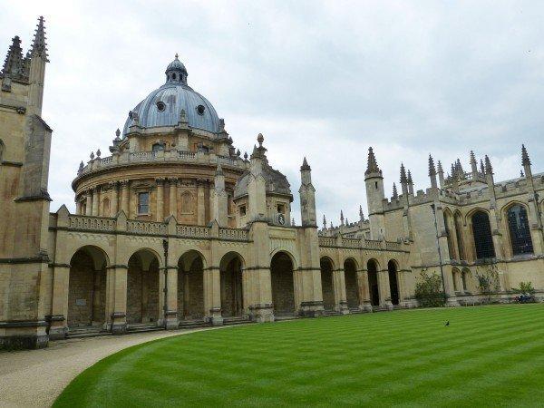 The inner courtyard of a venerable university consisting of a manicured lawn surrounded by a walking path which, in turn is ringed by limestone buildings. 