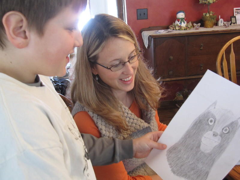 A woman in an orange top under a grey knit vest looks at a pencil drawing a child is showing her and smiling. 