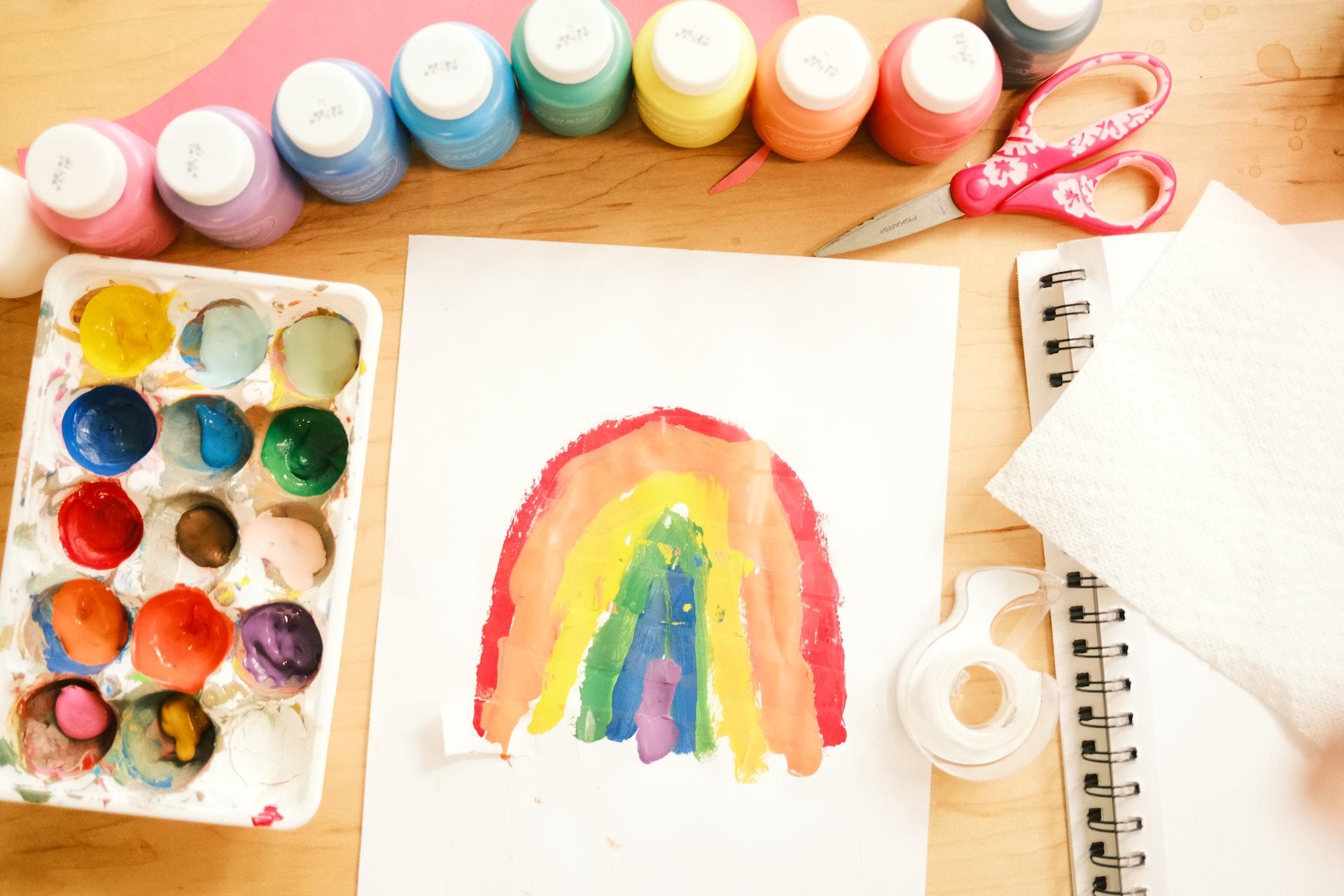 A child's rendering of a rainbow painted on a sheet of notebook paper, with an array of oil paint bottles lined up above the work and a tray of watercolours beside it