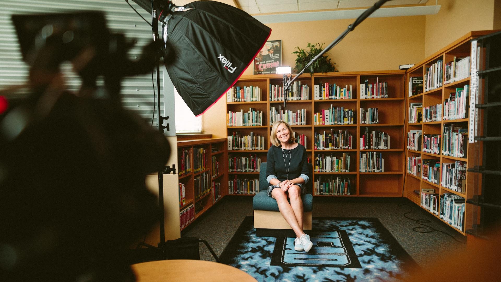 A blond woman in a dark jumper sits in a mock library with lights and cameras pointed at her and a microphone boom overhead