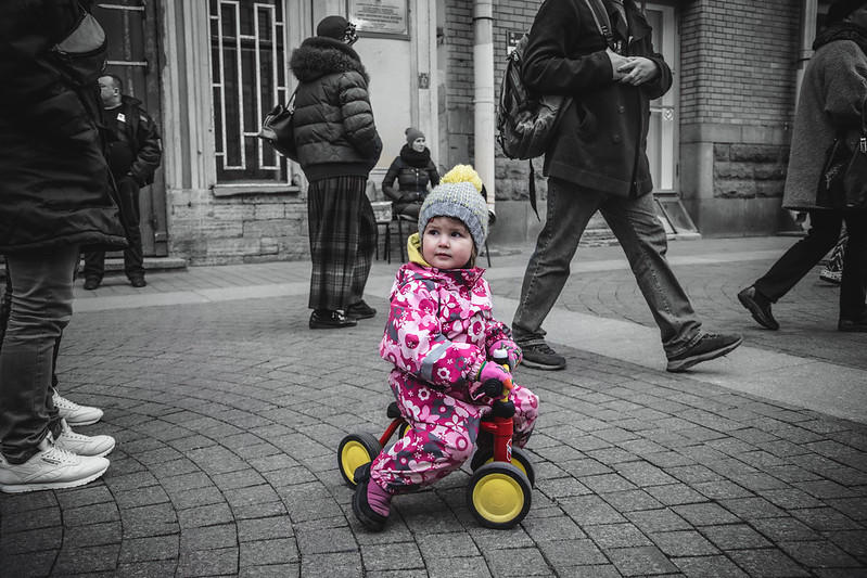 a small child dressed in a flowery pink outfit and wearing a grey hat sits atop a tricycle with yellow wheels while all around her people and buildings appear in greyscale. 