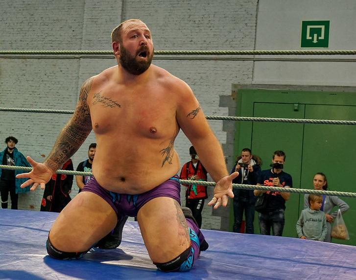 A wrestler wearing purple trunks an boasting a lot of tattoos kneels in the ring, shouting as spectators look on. 