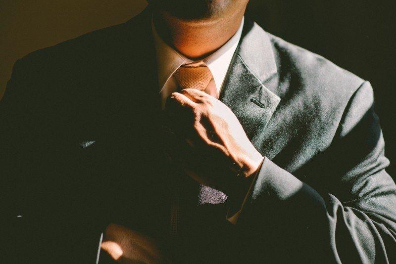 A person wearing a grey business suit is seen from the neck down as he stands in shadow, adjusting his orange-coloured tie with his left hand.