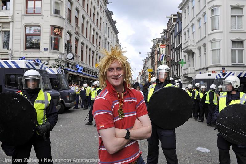 A young man in a fright wig wearing a red striped top stands in the street with his arm crossed in from of a line of riot police