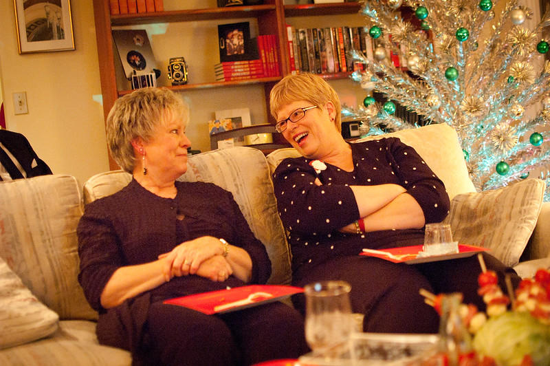 Two women sit side by side on a couch, looking at one another and smiling broadly while behind them, the silver and green Christmas tree gives off a festive air. 