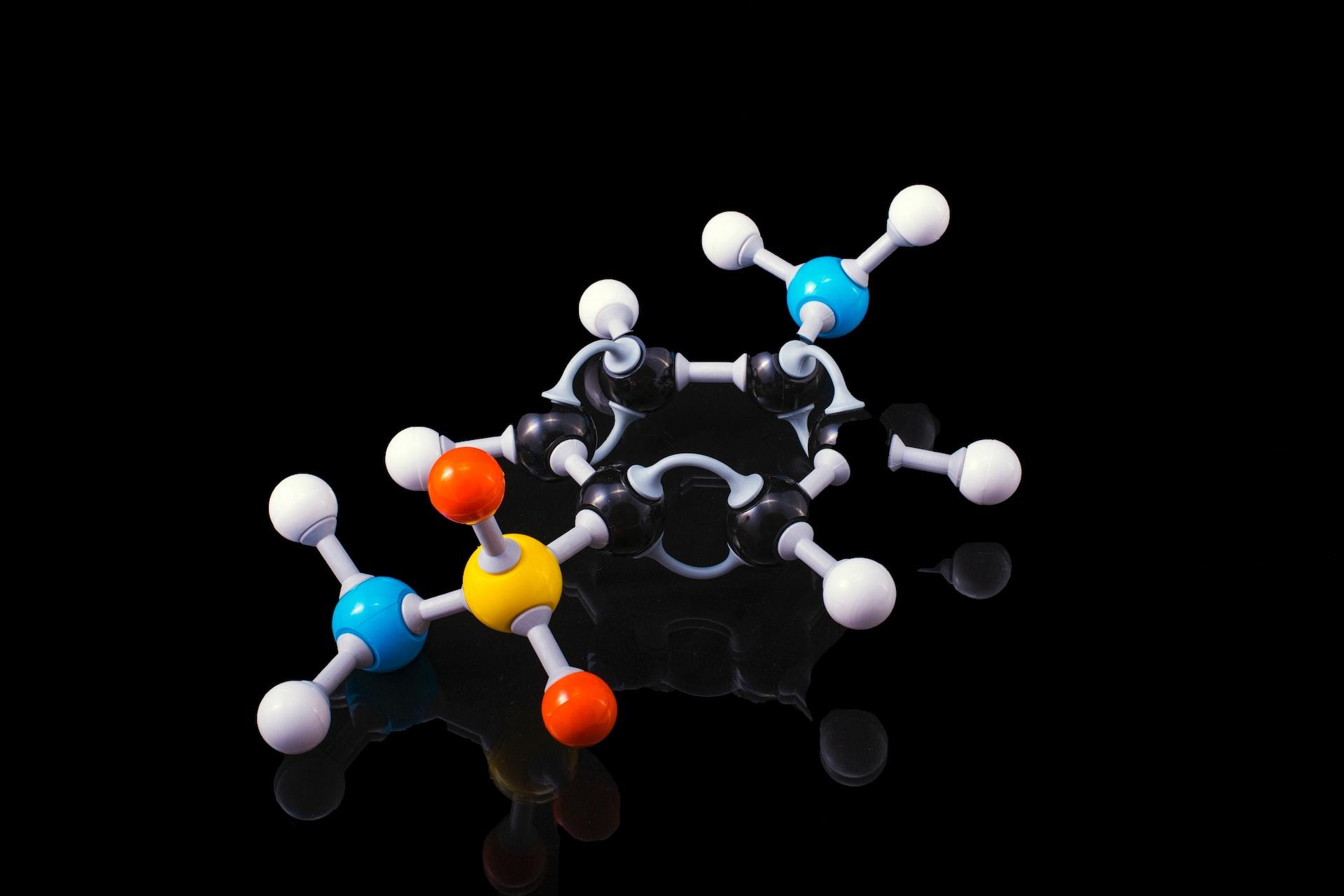 A sulfanilamide molecule made up of blue, red, grey, yellow and black balls against a black background. 