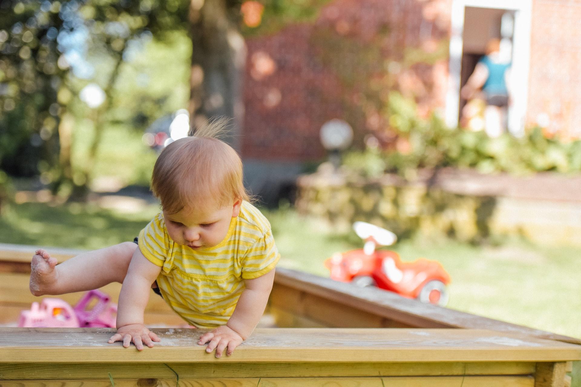 A toddler with blond hair in a yellow outfit climbs over the rim of a sandbox. 