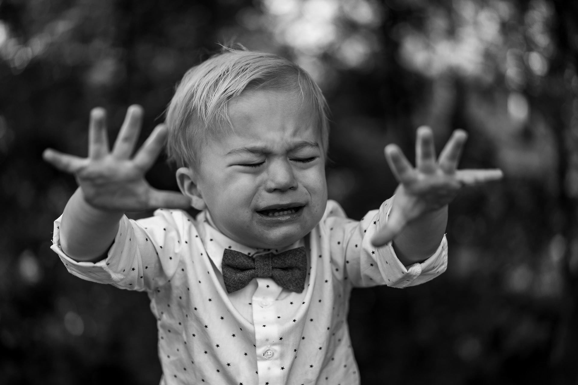 A black and white snap of a small boy wearing a dress shirt and bow tie with his hands up and fingers extended, crying.