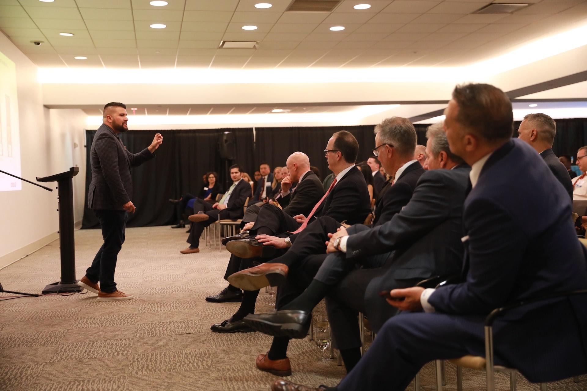 A man stands in front of a podium, addressing a crowd of seating men wearing business suits. 